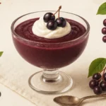 Glass bowl filled with rich purple chokecherry pudding, topped with whipped cream and fresh chokecherries, served with a spoon on a neutral background — chokecherry pudding recipe.